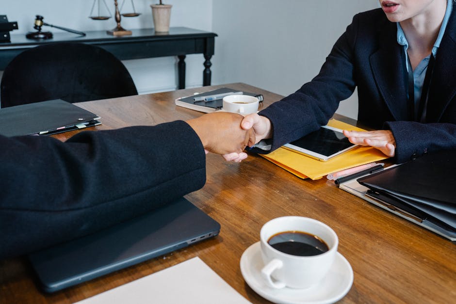 Professional business meeting handshake over a table with coffee, documents, and tech devices in an office setting.